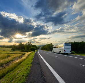 Ein weißer Reisebus fährt auf einer Landstraße in die Ferne, während die Sonne durch dramatische Wolken scheint.