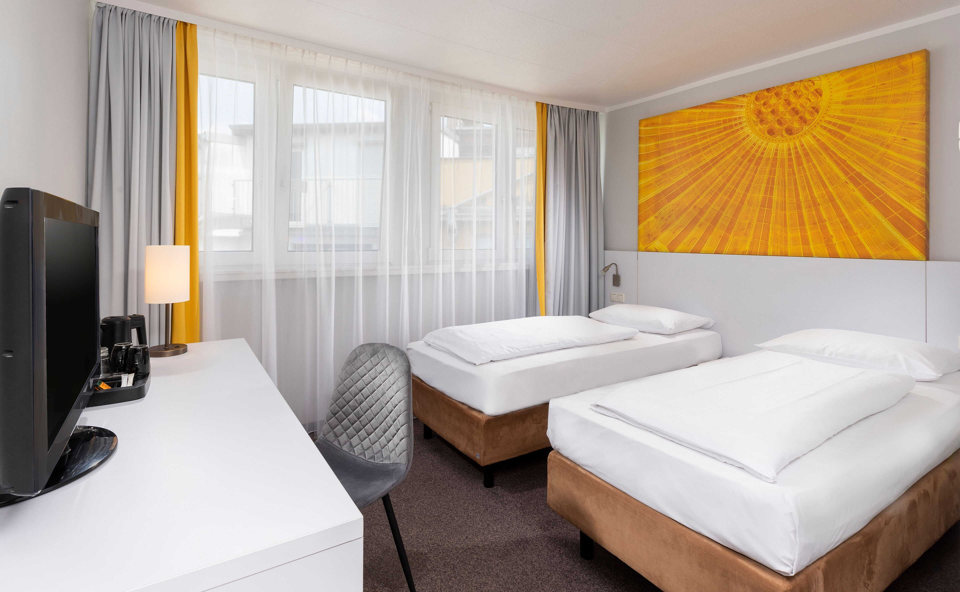 A modern hotel room features two neatly made twin beds, a vibrant yellow artwork, and a desk with a TV and lamp.