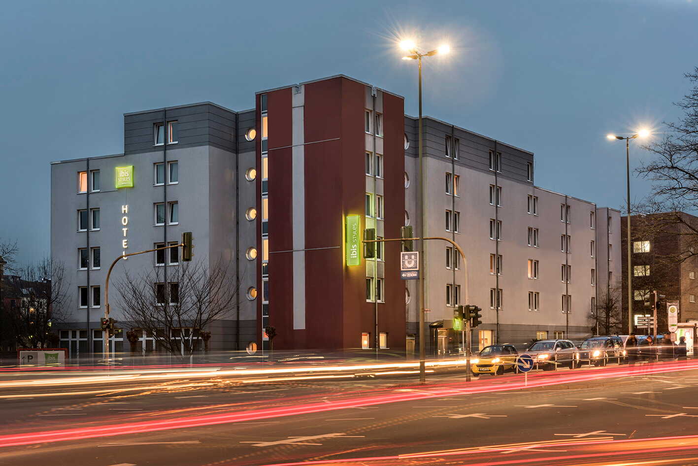 Cars drive past a modern hotel building at dusk, with bright streetlights illuminating the exterior.