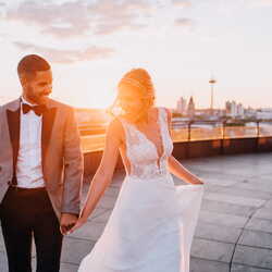 Ein elegantes Paar in festlicher Kleidung hält sich bei Sonnenuntergang auf einer Dachterrasse vor einer Stadtlandschaft an den Händen.
