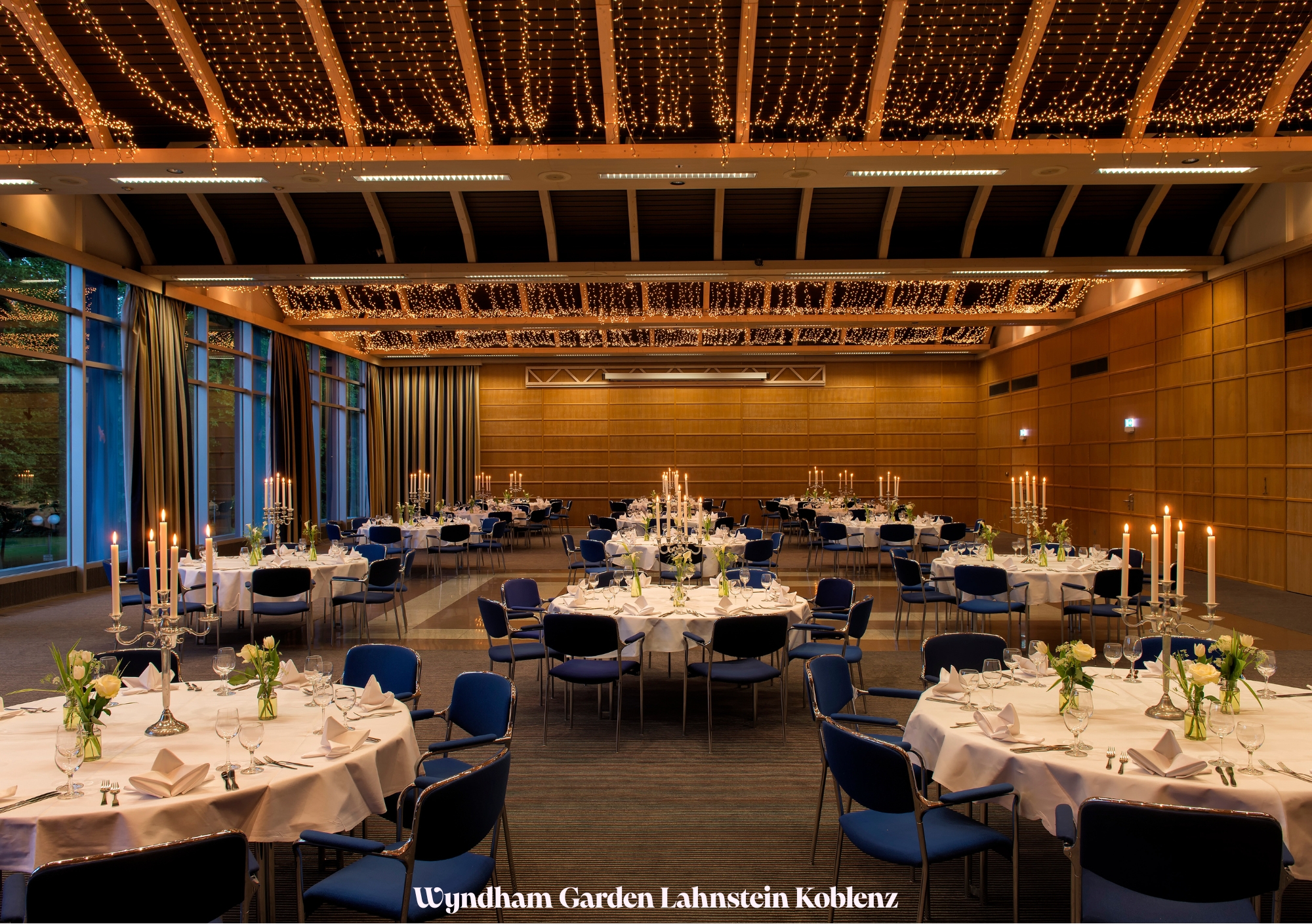 Ein elegant dekorierter Festsaal mit runden Tischen, Kerzenleuchtern und Lichterketten an der Decke.