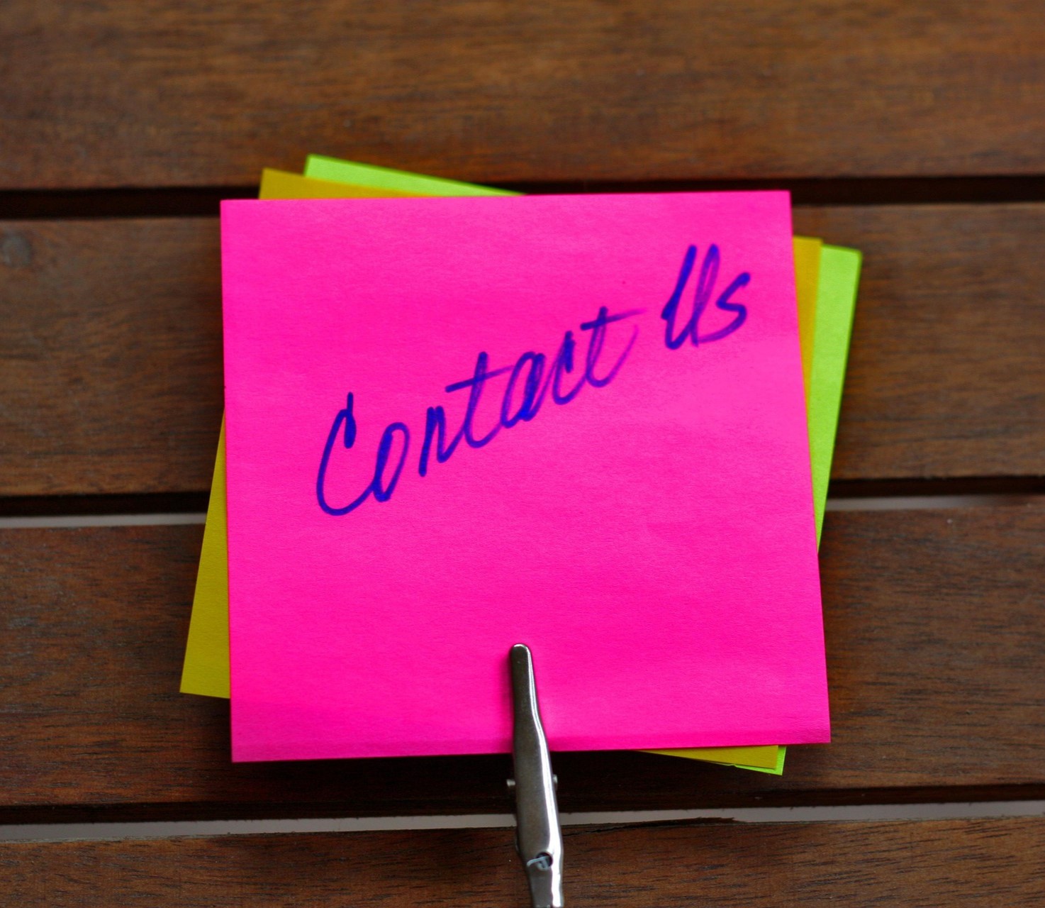 Ein pinker Zettel mit der Aufschrift "Contact Us" ist mit einer Klammer auf einem Holztisch befestigt.